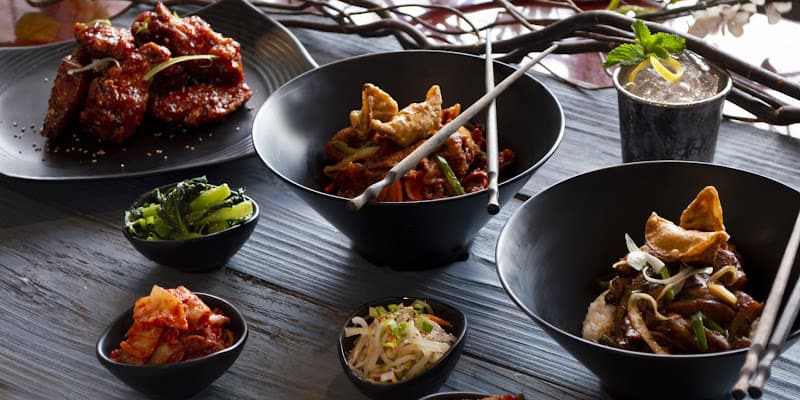 A full table at Matjoa House: Korean fried chicken wings, bowls, and a cocktail under pendant lighting